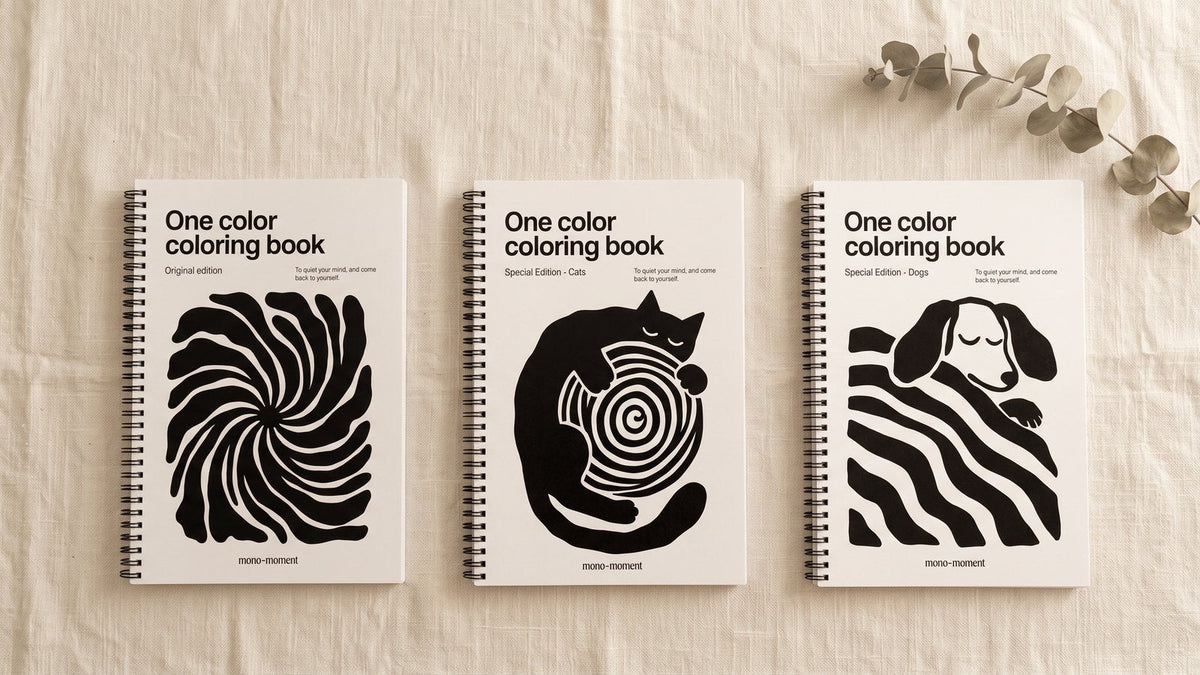 Three editions of the One Color Coloring Book laid side by side on a linen cloth