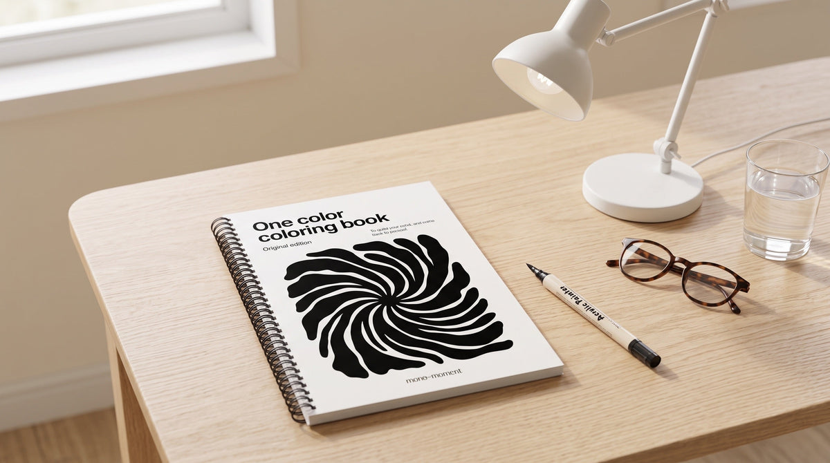 The One Color Coloring Book lying flat on a bright kitchen table beside a desk lamp, reading glasses and a brush pen