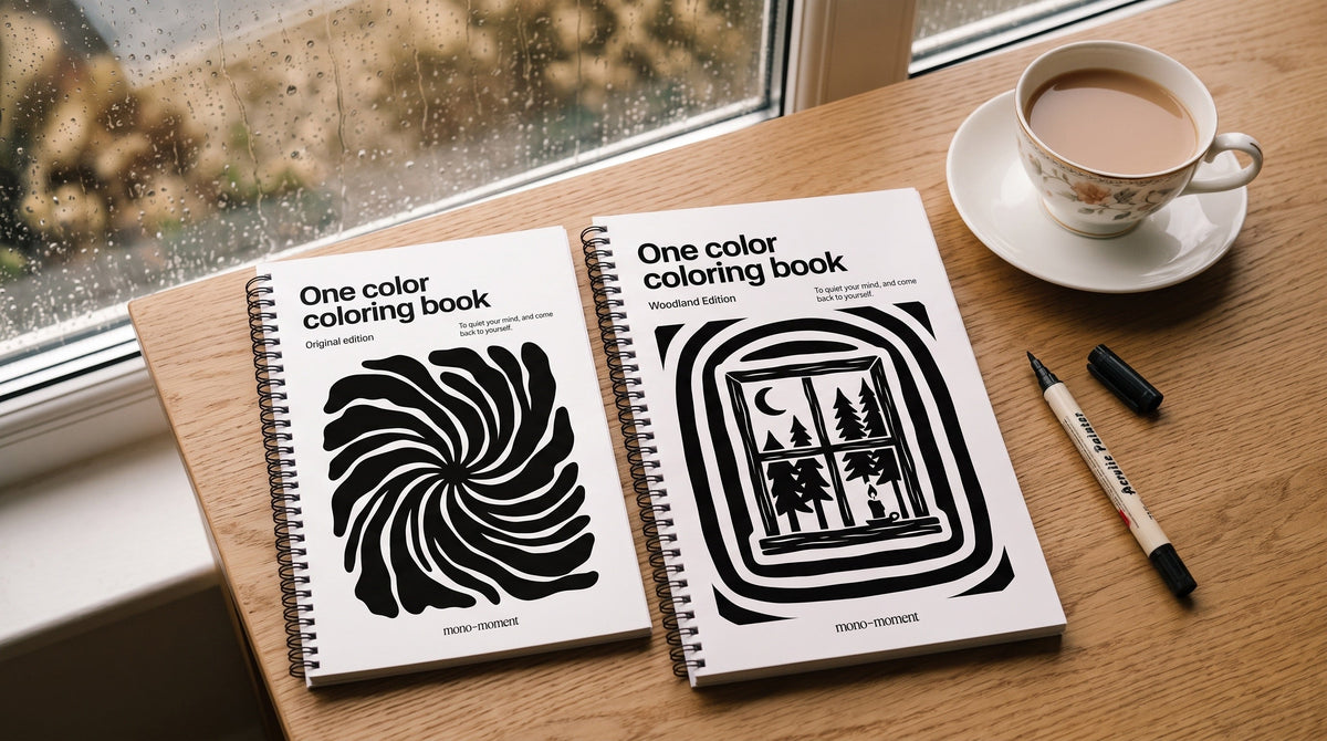 Two editions of the One Colour Colouring Book beside a cup of tea on a saucer by a rainy window