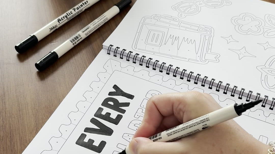 A hand coloring the bold letters of a quote page in the One Color Coloring Book, pens beside