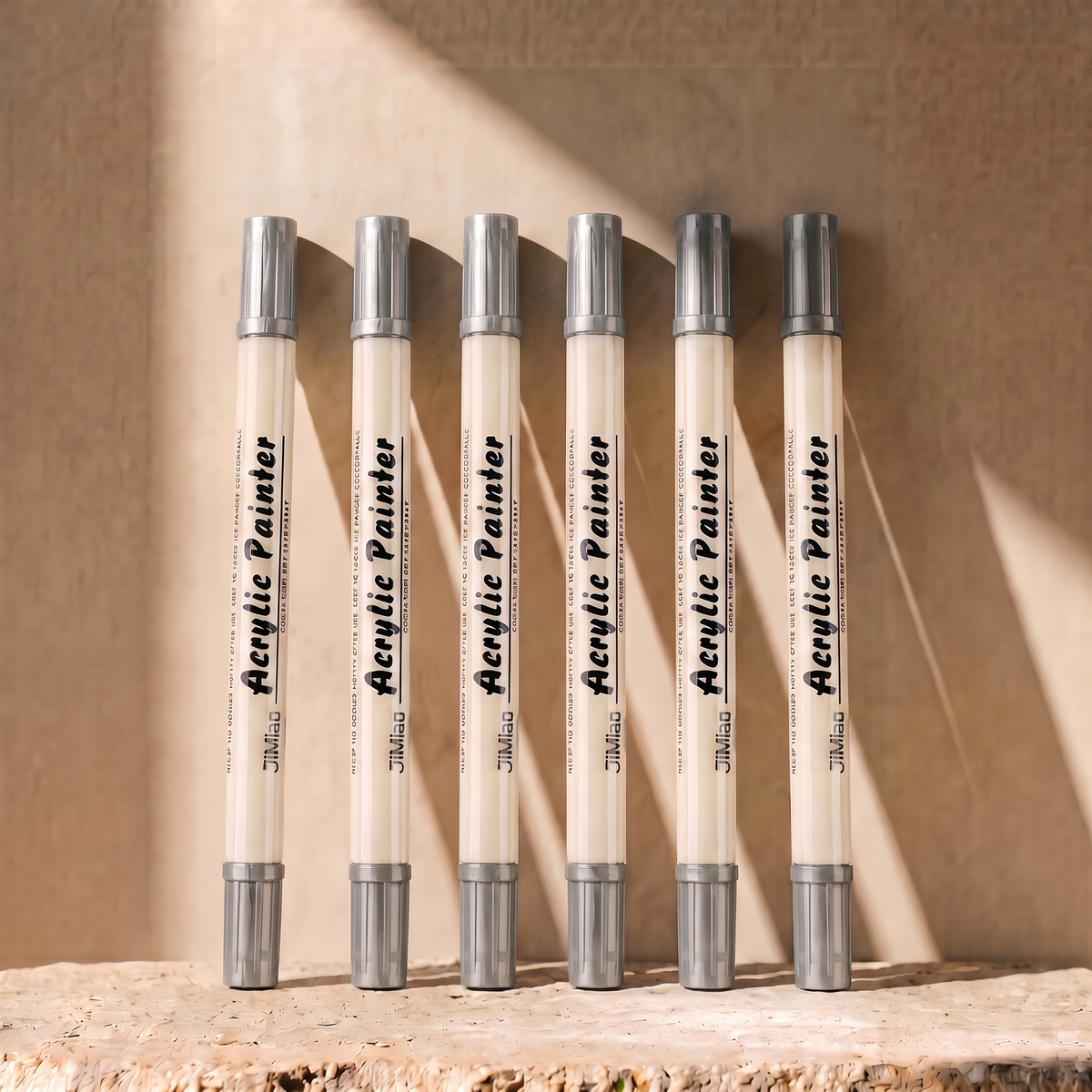 Six white acrylic paint markers with silver caps are lined up on a stone surface against a warm beige wall with dramatic sunlight and shadows.